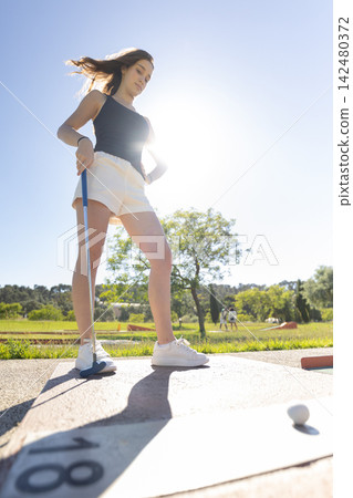 Girl practices her golf swing at the driving range during sunny afternoon in a green landscape with other players nearby Girl practices her golf swing at the driving range during sunny afternoon in a green landscape with other players nearby 142480372