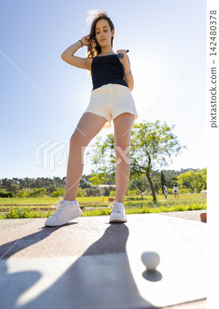 Girl prepares to hit golf ball during sunny day at the golf course with green landscape and blue sky Girl prepares to hit golf ball during sunny day at the golf course with green landscape and blue sky 142480378