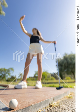 Woman plays mini golf on a sunny day in a park while taking a selfie with her phone near the green Woman plays mini golf on a sunny day in a park while taking a selfie with her phone near the green 142480419