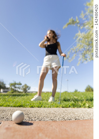 Woman plays golf on a sunny day in a park while talking on the phone and preparing to hit the ball 142480420