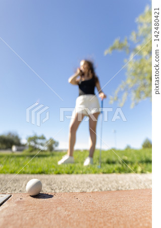 Woman golfing at outdoor park while focusing on small white ball on ground during bright sunny day Woman golfing at outdoor park while focusing on small white ball on ground during bright sunny day 142480421