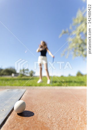 Woman plays golf in bright sunlight while an egg sits on the ground in the foreground at a park on a clear day Woman plays golf in bright sunlight while an egg sits on the ground in the foreground at a park on a clear day 142480422