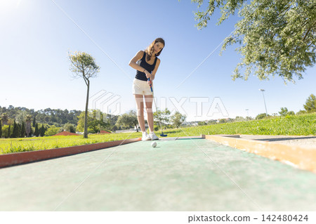 Mini golf game at a park on a sunny day with a player focused on putting the ball towards the hole Mini golf game at a park on a sunny day with a player focused on putting the ball towards the hole 142480424