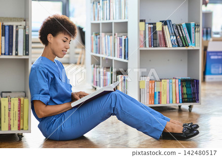 Dedicated medical resident absorbed in studying complex clinical guidelines from a thick manual between bookshelf rows Dedicated medical resident absorbed in studying complex clinical guidelines from a thick manual between bookshelf rows 142480517
