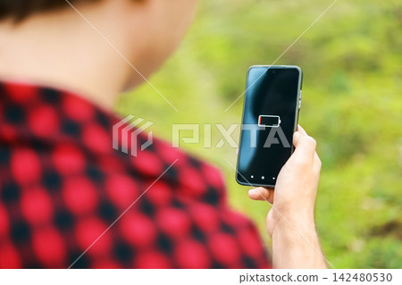 person holds a smartphone showing a low battery warning on screen, facing a battery emergency and power shortage while outdoors. person holds a smartphone showing a low battery warning on screen, facing a battery emergency and power shortage while outdoors. 142480530