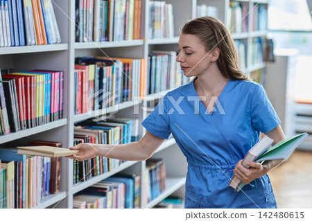 Caucasian female intern taking a textbook from a library shelf while holding study notes Caucasian female intern taking a textbook from a library shelf while holding study notes 142480615