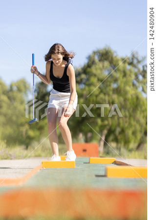 Young girl plays mini golf on outdoor course during sunny day in recreational area near trees 142480904