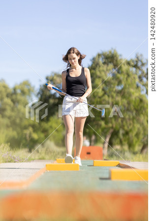 Young woman plays mini golf on sunny day in outdoor park setting with trees and clear blue sky Young woman plays mini golf on sunny day in outdoor park setting with trees and clear blue sky 142480920