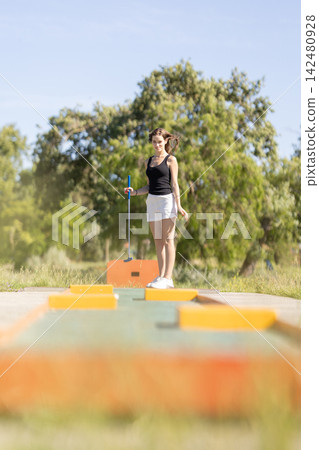 Woman playing mini golf on a sunny day in a park with trees and clear sky in the background Woman playing mini golf on a sunny day in a park with trees and clear sky in the background 142480928