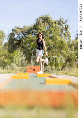 Girl plays mini golf on a sunny day in a park with green grass and trees around Girl plays mini golf on a sunny day in a park with green grass and trees around 142480931
