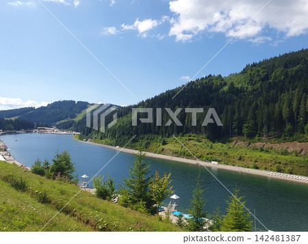 Scenic view of a mountain lake surrounded by dense green pine forest under a clear blue sky with white clouds on Scenic view of a mountain lake surrounded by dense green pine forest under a clear blue sky with white clouds on 142481387
