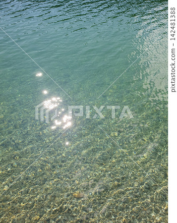 Clear turquoise water background with visible smooth pebble stone bottom in bright daylight. Sunlight sparkling on ripples Clear turquoise water background with visible smooth pebble stone bottom in bright daylight. Sunlight sparkling on ripples 142481388