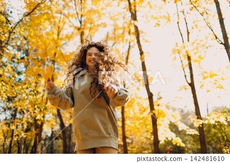 Smiling young woman plays and jumping dancing in the autumn forest with the yellow leaves at sunset. 142481610