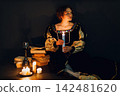 Young adult woman dressed in a medieval dress sitting near ancient books and candles with bottle of red wine and holding a glass of wine in darkness. 142481620