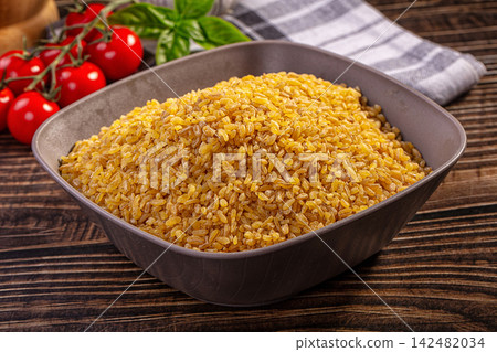 Raw bulgur wheat in a bowl with fresh cherry tomatoes and basil on a wooden table 142482034