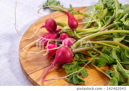 Fresh bunch of red radishes with green tops on round wooden cutting board Fresh bunch of red radishes with green tops on round wooden cutting board 142482043
