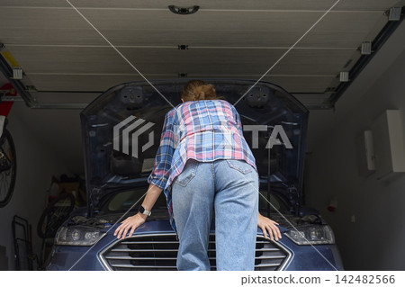 Woman leaning over front of car and looking under open hood in home garage Woman leaning over front of car and looking under open hood in home garage 142482566