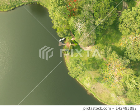 Aerial drone view of green lakeside park with small dock and boats on calm water. Nature travel, eco resort, waterfront leisure, and peaceful scenic landscape concepts. Eco tourism, wellness retreat. 142483882