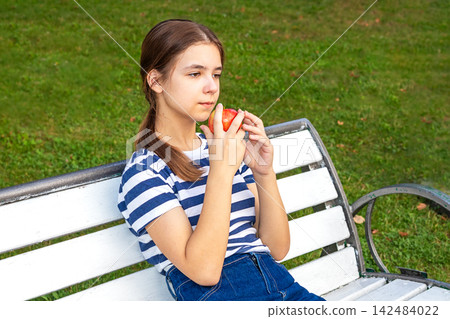 Girl eating an apple on a white bench in the park, thoughtful summer portrait Girl eating an apple on a white bench in the park, thoughtful summer portrait 142484022