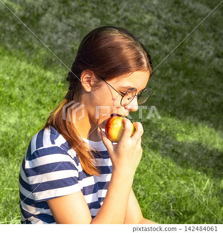 Girl with glasses eating a fresh apple while sitting on grass in sunny park 142484061