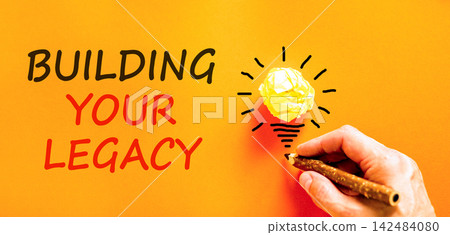 Building your legacy words symbol. Concept words Building your legacy on beautiful orange paper. Beautiful orange background. Businessman hand. Business building your legacy concept. Copy space. 142484080