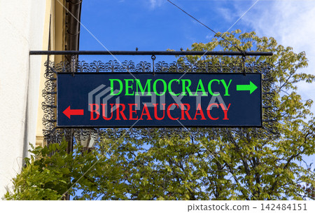 Democracy or bureaucracy words symbol. Concept words Democracy Bureaucracy on beautiful black road sign. Beautiful blue sky background. Business democracy bureaucracy concept. Copy space Democracy or bureaucracy words symbol. Concept words Democracy Bureaucracy on beautiful black road sign. Beautiful blue sky background. Business democracy bureaucracy concept. Copy space 142484151