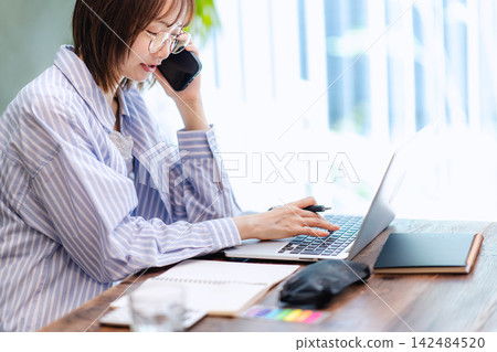 一位年輕女子一邊用筆記型電腦工作,一邊用智慧型手機打電話。 一位年輕女子一邊用筆記型電腦工作,一邊用智慧型手機打電話。 142484520