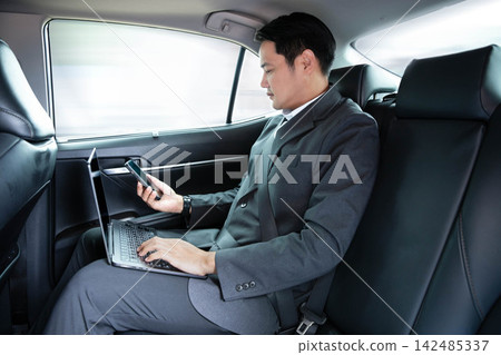 Asian businessman using laptop computer and holding smartphone while sitting in back seat of luxury car, working remotely during daily commute or business travel, reflecting corporate executive Asian businessman using laptop computer and holding smartphone while sitting in back seat of luxury car, working remotely during daily commute or business travel, reflecting corporate executive 142485337