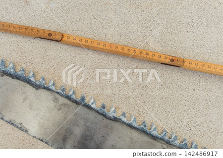 Durable old saw blade and a wooden folding ruler laying on a light textured concrete background, representing manual work, measurement, and traditional craftsmanship Durable old saw blade and a wooden folding ruler laying on a light textured concrete background, representing manual work, measurement, and traditional craftsmanship 142486917
