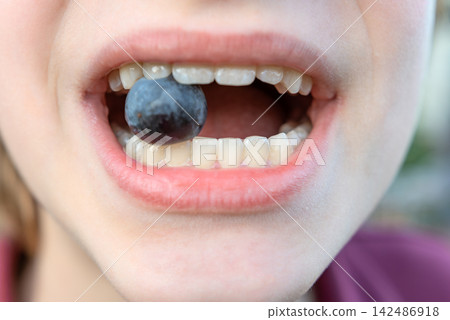 Young person playfully holding a fresh blueberry between their white teeth, representing concepts of childhood nutrition, oral health, and a healthy diet 142486918