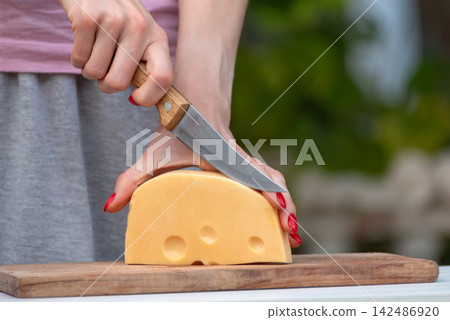 Woman's hands with red nails slicing a large block of yellow Swiss style cheese with a small knife on a wooden cutting board, close up food preparation scene Woman's hands with red nails slicing a large block of yellow Swiss style cheese with a small knife on a wooden cutting board, close up food preparation scene 142486920