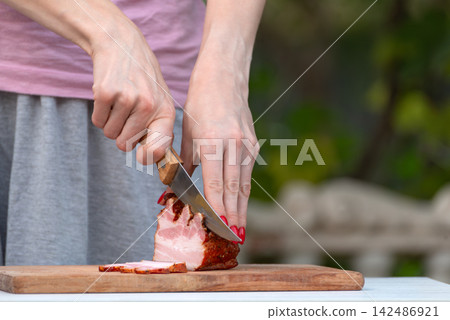 Woman's hands with red nails slicing smoked bacon on a rustic wooden cutting board outdoors, preparing homemade charcuterie style cold cuts for a gourmet snack or meal Woman's hands with red nails slicing smoked bacon on a rustic wooden cutting board outdoors, preparing homemade charcuterie style cold cuts for a gourmet snack or meal 142486921