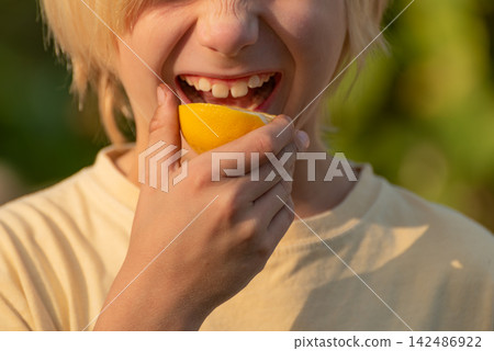 Child making a dramatic sour face while biting into a bright yellow lemon, playful reaction to intense citrus taste captured in a close up outdoor moment Child making a dramatic sour face while biting into a bright yellow lemon, playful reaction to intense citrus taste captured in a close up outdoor moment 142486922