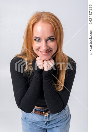 Young smiling woman with red hair and green eyes portraying happiness and a cheerful attitude, posing with hands clasped together on a light studio background Young smiling woman with red hair and green eyes portraying happiness and a cheerful attitude, posing with hands clasped together on a light studio background 142487136