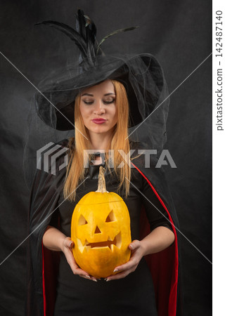 Young woman in witch hat and black cape holding a traditional carved jack o' lantern, celebrating the autumn spooky holiday season Young woman in witch hat and black cape holding a traditional carved jack o' lantern, celebrating the autumn spooky holiday season 142487140
