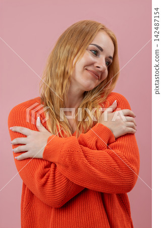 Young woman with blonde hair hugging her arms, projecting self love, acceptance, and comfort while smiling gently and looking to the side against a pink background Young woman with blonde hair hugging her arms, projecting self love, acceptance, and comfort while smiling gently and looking to the side against a pink background 142487154