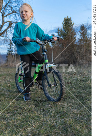Young blonde girl with a green and black bike stands on a grassy hillside, gazing ahead and ready for outdoor play and learning to ride, joyful and adventurous Young blonde girl with a green and black bike stands on a grassy hillside, gazing ahead and ready for outdoor play and learning to ride, joyful and adventurous 142487213
