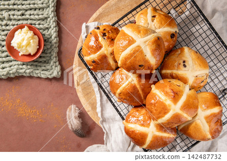Freshly baked golden hot cross buns sit on a cooling rack over a wooden board accompanied by a bowl of butter and decorative feathers on a warm pink surface for a cozy festive food scene Freshly baked golden hot cross buns sit on a cooling rack over a wooden board accompanied by a bowl of butter and decorative feathers on a warm pink surface for a cozy festive food scene 142487732