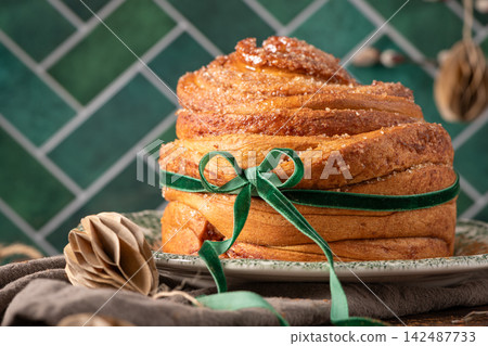 A close-up of a beautifully baked puff pastry Easter cruffin coated in cinnamon sugar and tied with a lush green velvet ribbon against a stylish green tiled backdrop A close-up of a beautifully baked puff pastry Easter cruffin coated in cinnamon sugar and tied with a lush green velvet ribbon against a stylish green tiled backdrop 142487733