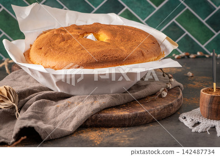 A beautiful Pao de Lo sponge cake wrapped in white parchment paper sits on a rustic wooden board against a vibrant green tiled wall with cozy kitchen decor 142487734
