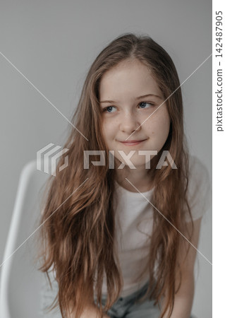 Preteen girl with long hair looking away while sitting on white chair in studio Preteen girl with long hair looking away while sitting on white chair in studio 142487905