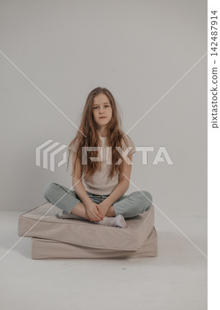 Preteen girl with long hair sitting cross legged on floor cushions against white background Preteen girl with long hair sitting cross legged on floor cushions against white background 142487914