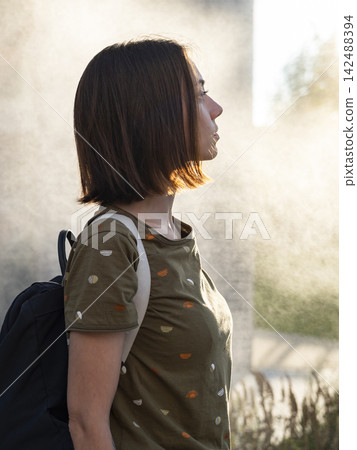 Young woman standing in front of a cooling fog system, illustrating summer lifestyle adaptations and comfort in warm weather. 142488394