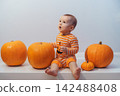 Halloween background with little surprised Caucasian baby in orange costume with lantern jack sitting among pumpkins on white background. Autumn fall harvest festival concept. Copy space 142488408
