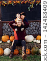 Happy family joyfully sitting on hay with pumpkins on pumpkin patch. Selecting best pumpkins for Thanksgiving and Halloween holidays decoration on farm. Pumpkin harvest. Autumn fall festive mood 142488409
