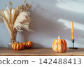 Cozy warm autumn home interior - decorative orange ceramic pumpkins, candles, dried flowers on the wooden console with white wall background. Scandinavian minimalist hygge home fall decor 142488413