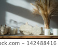 Cozy warm autumn home interior - white knitted pumpkins, wooden statuette, candles, dried flowers on the wooden console with white wall background. Scandinavian minimalist hygge home fall decor. 142488416