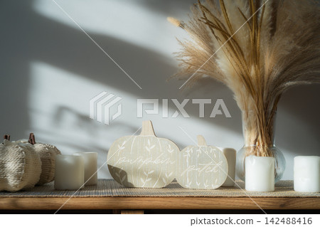 Cozy warm autumn home interior - white knitted pumpkins, wooden statuette, candles, dried flowers on the wooden console with white wall background. Scandinavian minimalist hygge home fall decor. Cozy warm autumn home interior - white knitted pumpkins, wooden statuette, candles, dried flowers on the wooden console with white wall background. Scandinavian minimalist hygge home fall decor. 142488416