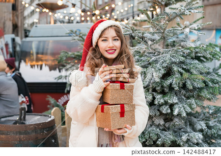 Young woman in Santa hat with with a tower of gift boxes on traditional city street Christmas market. Happy woman choosing Christmas gifts. Merry Xmas Happy New Year festive shopping, sales, discount. Young woman in Santa hat with with a tower of gift boxes on traditional city street Christmas market. Happy woman choosing Christmas gifts. Merry Xmas Happy New Year festive shopping, sales, discount. 142488417