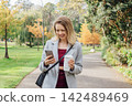 Smiling young woman using her phone while walking in autumn park for chatting, shopping, remote work, content creation for social media. Girl holding reusable cup with hot drink. and checking massages 142489469